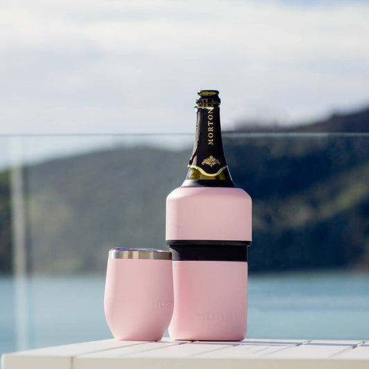 Pink bottle cooler with a bottle and two pink glasses on a railing with a scenic background.